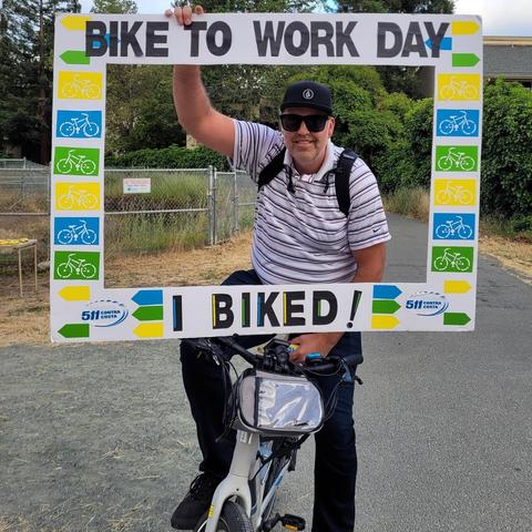Photo of a man on a bicycle holding up an oversized I Biked to Work photo frame which makes him look like he's inside the frame for this picture.