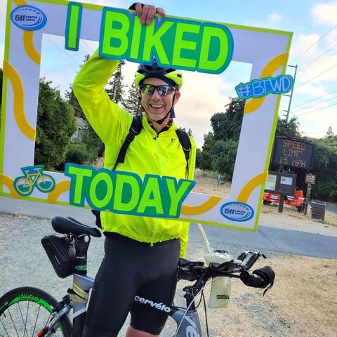 Photo of a man on a bicycle with a yellow jacket and sunglasses holding up an oversized I Biked to Work photo frame which makes him look like he's inside the frame for this picture.