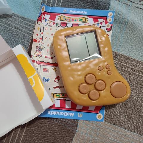 Photograph of a plastic framed Chicken McNugget toy with an LCD screen and buttons on it. Beneath it is a McDonald's sticker sheet. This is all sitting on a blue and grey tablecloth.