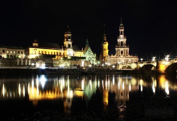 Dresdner Altstadt-Skyline bei Nacht
Die Gebäude sind in unterschiedlichen Lichtern angeleuchtet, rechts eine Brücke, die Lichter sind sternenförmig.
Die Altstadt spiegelt sich in der vordergründigen Elbe durch die Langzeitbelichtung gut ab.