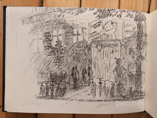 Black Fineliner on paper.

A big entrance of a house at night.
 On the boardwalk are many bycicles and some people are standing in front of the entrance. A big figure is standing at the side of the entrance. Light comes from the door and the windows. Spot lights above the entrance light a sign with a raised fist and the text "tuntenhaus bleibt". Many posters are on the wall, some have text on it. Garbage and stuff is lying around, garbage containers are there too. Trees and plants are on the house and in front of it.