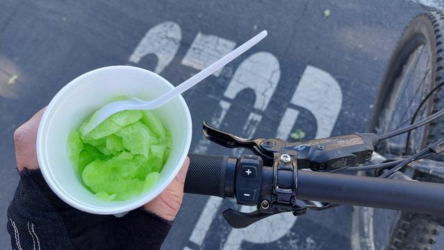 Photo of a gloved-hand holding a white dish with green-colored ice.  The hand and dish are resting on the handlebar of an eBike and part of the word STOP is visible on the pavement.