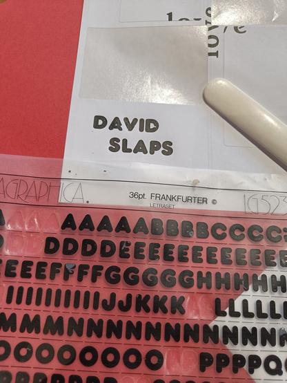 Worktable with a partially used sheet of Frankfurter 36pt (a well rounded sans serif font) sheet of Letragraphica rub-down lettering, and a sticker I made with it that says "DAVID SLAPS".