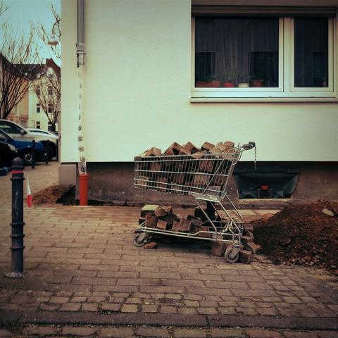 deutsch: ein quadratisches bild zeigt einen einkaufswagen, der gänzlich gefüllt ist mit pflastersteinen. der einkaufswagen steht an einer straßenecke vor einem weißen haus. offenbar wird um das haus herum am boden bzw. an den wänden gearbeitet, und die steine werden im wagen zwischengelagert. rechts neben dem einkaufswagen ist ein haufen erde. rechts oben ist noch ein fenster des hauses zu sehen.

english: a square picture shows a shopping trolley completely filled with cobblestones. the trolley is parked on a street corner in front of a white house. apparently, work is being carried out on the floor or walls around the house, and the stones are being temporarily stored in the trolley. to the right of the trolley is a pile of earth. a window of the house can still be seen at the top right.
