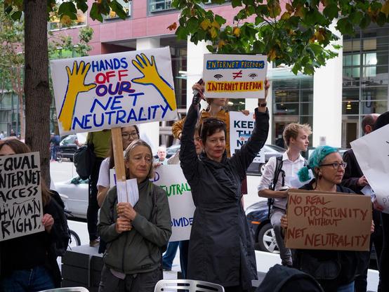 Protestors in San Francisco hold signs on sidewalk that read "Hands Off Our Net Neutrality" and "Internet Freedom does not equal Internet Fiefdom." Another sign reads: "Equal Internet Access is a Human Right"