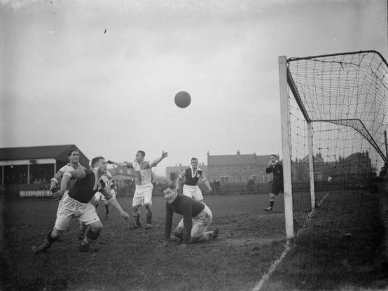 Eine Strafraumszene un FA Amateur Cup 1938 zwischen PALT Erith and Belvedere und Worthing. Ein Team trägt dunkle Trikots und weiße Hosen, das andere zweifarbig karierte Trikots und weiße Hosen. Ein Spieler im karierten Trikot hebt beide Arme und schaut zur Seitenlinie. Ein Spieler in dunklem Trikot schaut auf den Ball in der Luft, und plant, ihn gleich am gegnerischen Torhüter (bereits geschlagen am Boden) ins Tor zu schießen. Doch der Schiedsrichter pfeift im Hintergrund gerade. Ob es einen Elfmeter gab? Credit: IMAGO/TopFoto