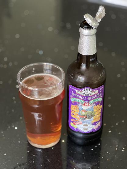 A pint glass of amber colored beer next to a 550ml bottle with a silver foiled cap and a label in many colors of green yellow reds and purple. In the center is a train with a red bow on it
