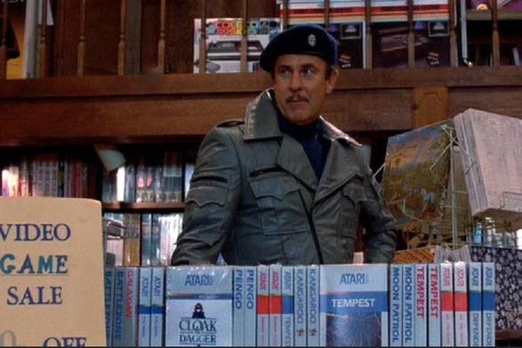 A movie still: A man wearing a military-style jacket and beret stands behind a display of Atari video game boxes in a store. A sign next to him reads, "VIDEO GAME SALE."