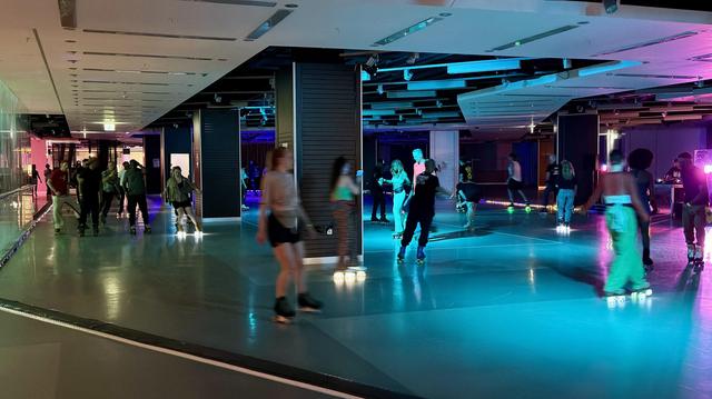 Menschen, die auf einer Indoor-Eisbahn mit bunter Beleuchtung und einem DJ im Hintergrund Rollschuhlaufen.