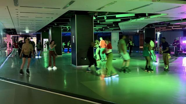 Menschen, die auf einer Indoor-Eisbahn mit bunter Beleuchtung und einem DJ im Hintergrund Rollschuhlaufen.