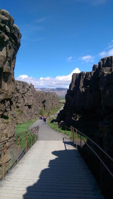 A path between rocky cliffs of Þingvellir with people walking in the distance, under a clear blue sky.