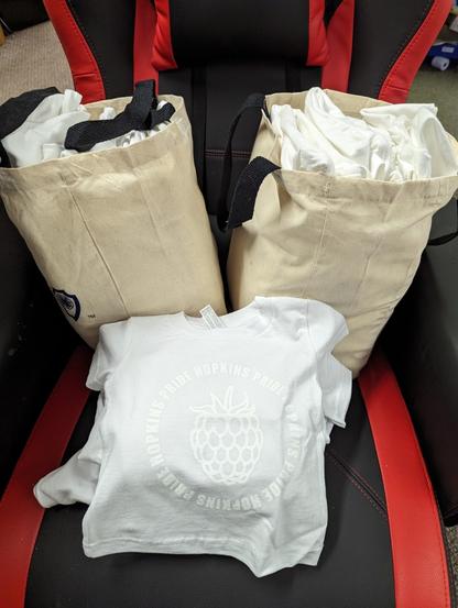 A photo of a lot of shirts on a chair. There are two linen-colored bags full of adult sizes & a stack of kid sizes.

The shirts are white with white printing. The phrase "Hopkins Pride" encircles a giant raspberry.