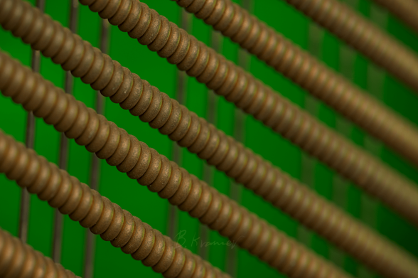 A macro photo of large copper-colored piano wires, stretching diagonally across the frame. Other sets of wires run vertically in the background, and all are lit with a green light from behind.