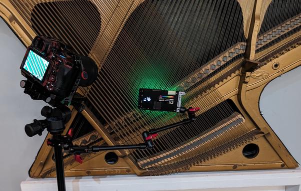A piano "plate" with strings installed, mounted on a wall. In front of it is a camera on a tripod, set at a close tilted angle. A mounting arm is attached to the tripod and has a pocket LED mounted on it, shining green light through the strings to bounce light off the background.
