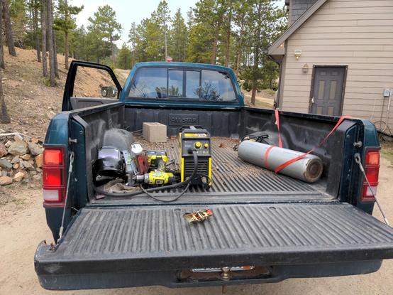 A 1994 Dodge Dakota, shot from the back with the tailgate down. The bed contains a welder, some hand tools and protective gear, and a tank of welding gas (don't worry, it was fully strapped in before I left). In the background, a garage is partially visible to the left, and trees take up most of the area in front of the truck.