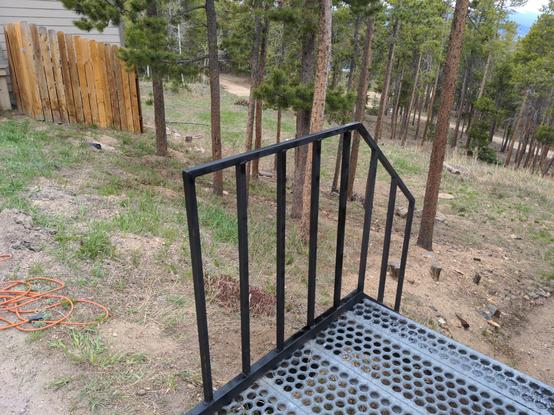 A metal railing and staircase. The railing has been recently rewelded and painted with fresh black paint. In the background are some scattered pine trees and an open field (septic field). A section of fence and part of the garage building are visible.