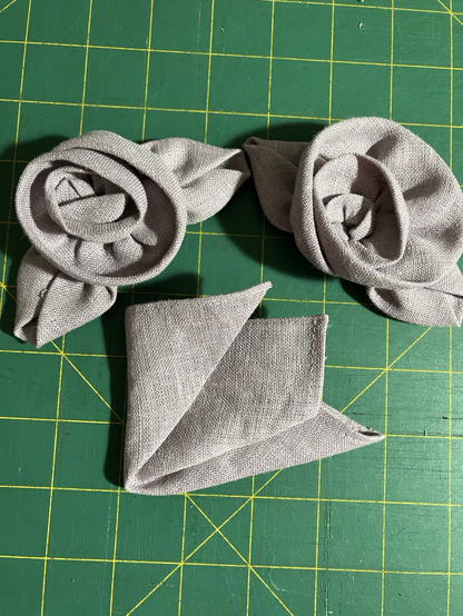 Two linen fabric roses and a triple folded matching pocket square on a green cutting mat