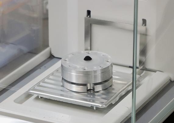 In der Bildmitte befindet sich auf einem runden Metallkörper ein kleines Häufchen tiefschwarzes Pulver.
Das ganze befindet sich in einer von Glas umschlossenen Feinwaage.
(Bild: Rigaku via Business Wire)