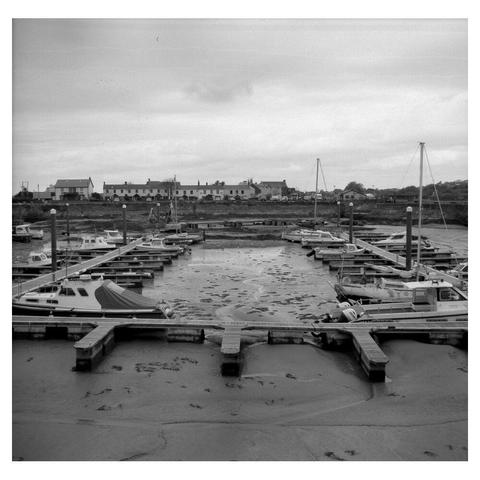 a black and white photo of boats lined along a square pontoon. The tide is out and the harbour floor is muddy