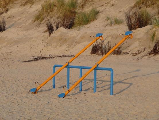 Am Strand steht eine Wippe, im Hintergrund steigt die Düne auf. Genau genommen sind es zwei Wippen, die so parallel stehen, als ob sie das Bild in seiner Diagonalen teilen. Die satt-gelben Wippen mit ihren hellblauen Sitztellern sind an einem großen, ebenfalls hellblauen Bügel befestigt, dessen Fundament im Sand eingegraben ist. Das abendliche Sonnenlicht feuert die Farben nochmal an und eirzeugt eine warme Stimmung.

There is a seesaw on the beach, with the dune rising in the background. Strictly speaking, there are two seesaws standing parallel to each other as if they were dividing the picture diagonally. The rich yellow seesaws with their light blue seat plates are attached to a large, light blue bracket, the base of which is dug into the sand. The evening sunlight fires up the colors once again and creates a warm atmosphere.