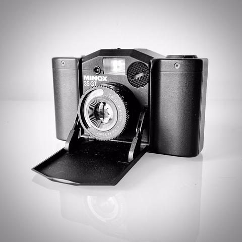 deutsch: quadratisches schwarzweißbild zeigt die analoge kleinbildkamera minox 35gt mit geöffneter objektivklappe. sie steht auf weißem spiegelndem glas und vor einer weißen wand.

english: square black and white picture shows the analogue small format camera minox 35gt with the lens cap open. it stands on white reflective glass and in front of a white wall.