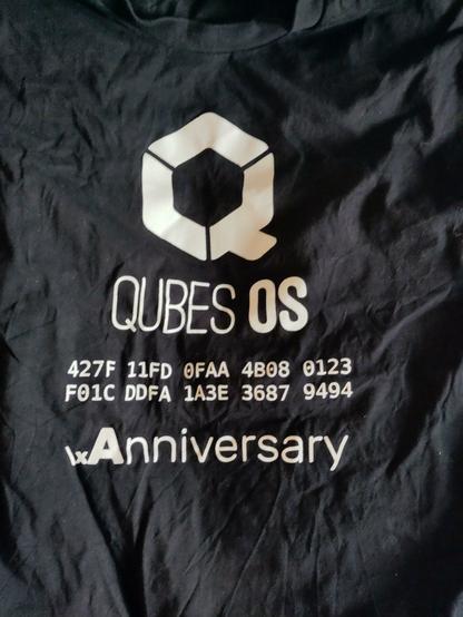 Photo of a somewhat crumpled black t-shirt with white QubesOS logo and white text:

Qubes OS

427F 11FD 0FAA 4B08 0123
F01C DDFA 1A3E 3687 9494

\xAnniversary