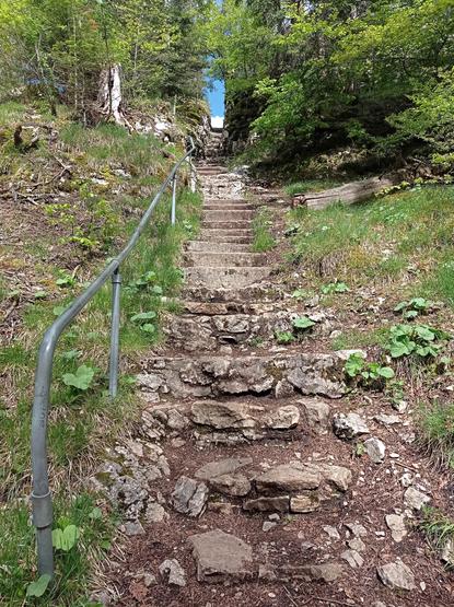 Vue depuis le bas d'un long escalier.
À droite, un rampe métallique.
Les marches sont d'abord en cailloux, puis en béton (très ancien), puis taillées dans le rocher.
Tout en haut, l'escalier semble déboucher sur le ciel tout bleu.