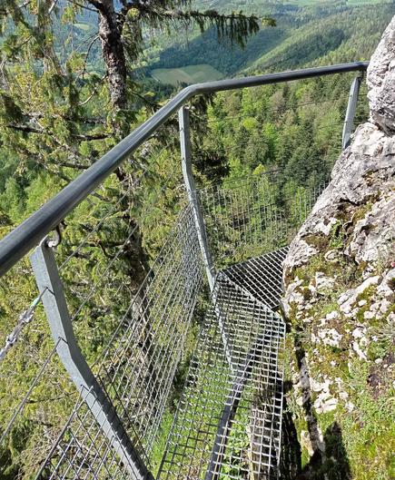 Une passerelle étroite contourne un rocher au-dessus du vide. Garde-fou solide !
En contrebas dans le lointain, on devine des collines boisées et un champ.