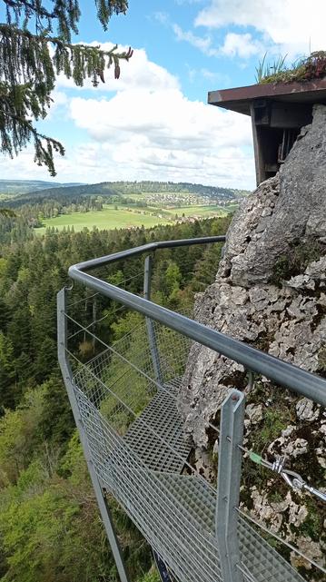 La même passerelle et le sommet d'un bâtiment qui dépasse en haut du rocher à droite.
Au loin, un paysage de colline boisée au pied de laquelle est niché un village, et des champs.
Ciel bleu et nuageux.
