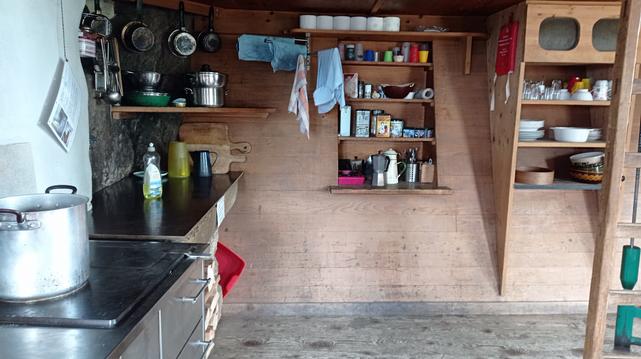 Intérieur du refuge.
Solide cuisinière sur laquelle est posé un gros faitout, plan de travail, rangements contenant ustensiles de cuisine, vaisselle, torchons, ingrédients non périssables...
La paroi du fond est inclinée selon la forme du rocher.