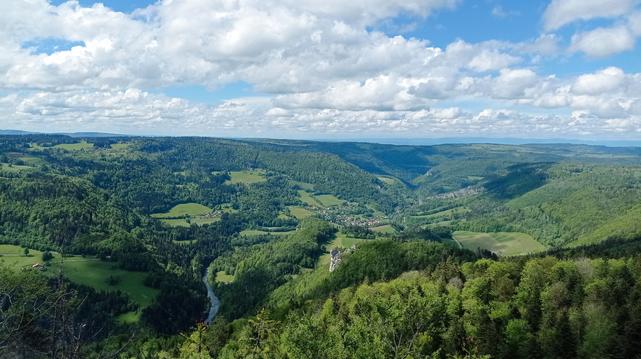 Vue sur une vallée au fond de laquelle coule une rivière.
Dans le fond de la vallée, un village et une barre de rocher qui s'avance pour la dominer.
En arrière plan, l'aval de la vallée, entre des collines boisées.
En dernier plan à l'horizon, une chaîne de montagnes moyennes.
Ciel nuageux mais clair.
