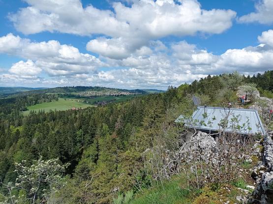 Par rapport à la photo précédente, vue à l'opposé géographique.
À droite, le tout zingué du refuge, des rochers et des randonneurs.
À gauche, ouverture sur le même paysage que depuis la passerelle : forêt, champs, colline et village.
Ciel bleu et nuageux.