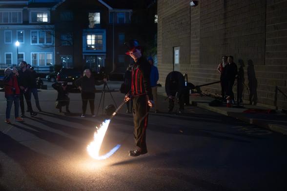 A street performer in a top hat and red-lined black outfit skips rope with a flaming whip in front of an audience at night.