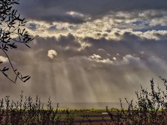 View of the sky in a brake from the rain. There's an opening between the clouds, the sunlight is coming in drawing rays of light in the sky. In the lower part a countryside is lit in bright green.