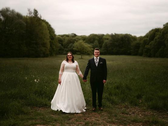 In the photo, a bride and groom stand side by side, holding hands, in the middle of a green field. The bride is wearing a long white wedding dress with lace sleeves and a V-neckline, while the groom is dressed in a dark suit with a white shirt and patterned tie. The background consists of lush greenery and trees, with a slightly overcast sky above, giving the scene a calm and serene atmosphere. The couple looks directly at the camera, standing still and composed, with the natural setting emphasizing a sense of intimacy and tranquility. This beautiful moment was captured by the Hamburg elopement and wedding photographer Sturmsucht.