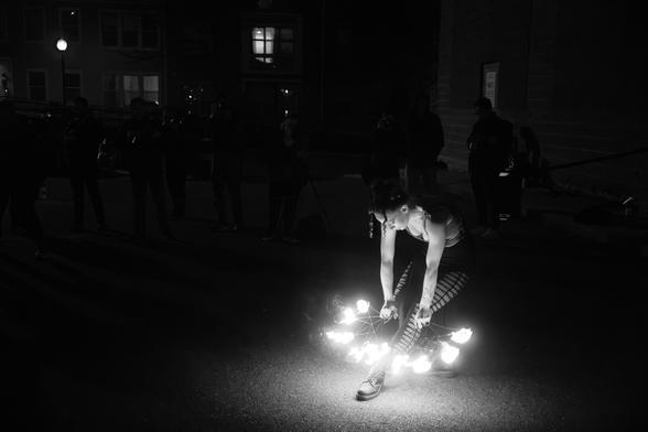Black and white photo of a fire dancer performing at night, with an audience in the background, surrounding their leg and foot with flames.