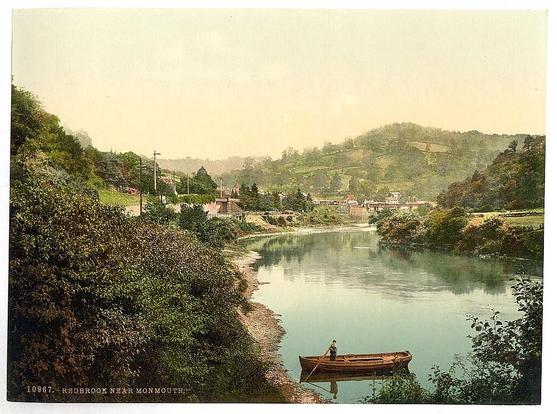 The image depicts a serene rural landscape. In the foreground, there's a calm body of water that resembles a lake or river. A small boat is visible on the left side of the waterway, suggesting some form of recreational activity or transportation.

The middle ground features lush greenery and what appears to be a wooden bridge or path crossing the waterway. This suggests the presence of human habitation in the vicinity. The surrounding hills are dotted with trees and houses, contributing to the idyllic countryside atmosphere.

In terms of alt text, this image captures the essence of rural life in a specific location between 1890 and 1900. It conveys tranquility and the beauty of nature, with man-made elements like the bridge or path indicating human interaction with the environment.