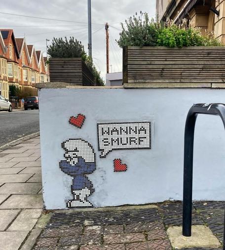 Streetartwall. A small, funny paste-up made of painted mosaic tiles has been stuck to a low street wall. A blue Smurf with a white pointed cap and trousers stands there and smiles. Above him is a speech bubble with the words "Wanna Smurf" and two red hearts next to it.
