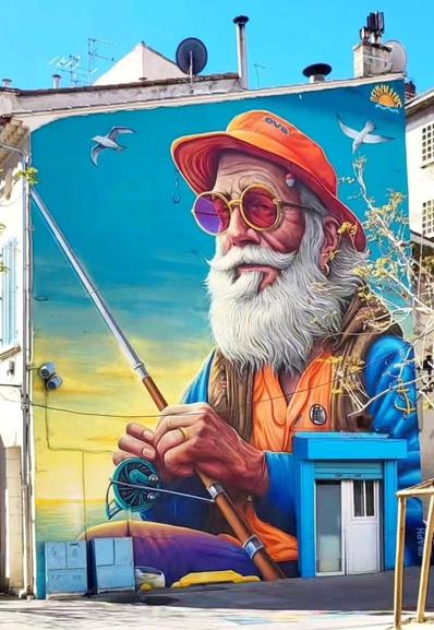 Streetartwall. A beautiful mural of an old sailor was sprayed/painted on the outside wall of a single-storey building with a wooden door. The old man with the long gray beard is wearing an orange hat and sunglasses with orange lenses. He is wearing an orange T-shirt and a beige vest without sleeves over a blue jacket. An anchor is embroidered on the vest and a fishing rod is tucked into the vest. He is holding a fishing rod in his hands and gazing contentedly into the distance. Seagulls are flying in the blue/green/yellow background, waiting for prey. A great mural in the middle of the pedestrian zone of the fishing village.