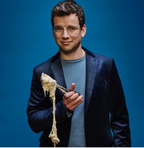 Pieter Derks staat op de foto met een lichtblauw shirt met daarover een donkerblauw colbert met een schep in zijn hand met oliebollenbeslag wat daaruit lekt.