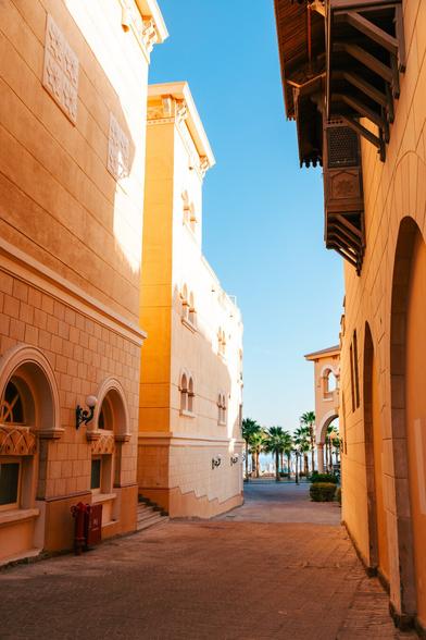 This image showcases a narrow alleyway in what appears to be a Mediterranean or Middle Eastern setting. The architecture includes two-story buildings with arched windows and doors, and the
facades are embellished with decorative elements such as masonry work and tiles. A clear blue sky overhead suggests fair weather conditions. There's no visible text in the image to provide
additional context.