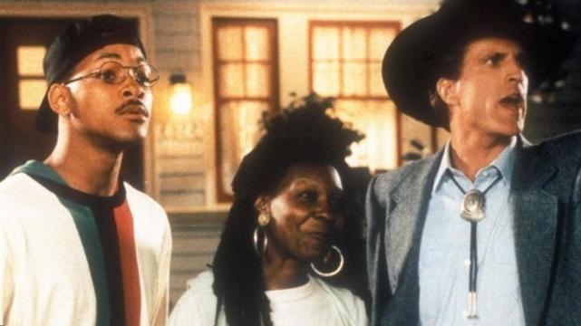 Will Smith (in mustache, round wire frame glasses and baseball cap), Whoopi Goldberg with big hoop earrings, and Ted Danson in a big cowboy hat and western tie, standing in front of a warmly lit home.