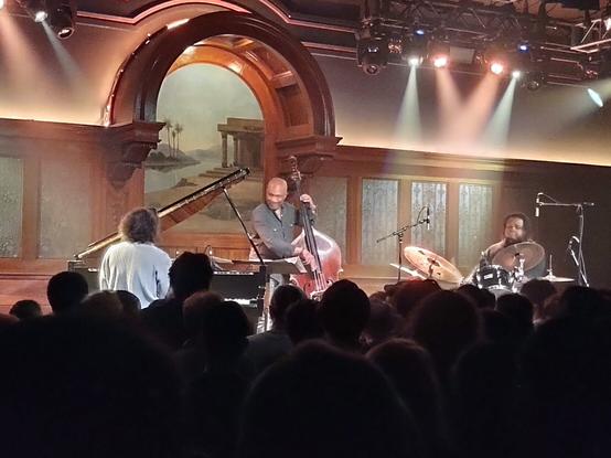 Emmet Cohen with a head of crazy hair sits at the piano on the left, Reuben Rogers on Upright bass in center and Kyle Poole on drums to the right. on the stage of the lodge room which is a converted 1922 Masonic Lodge location with arched arc alcove background