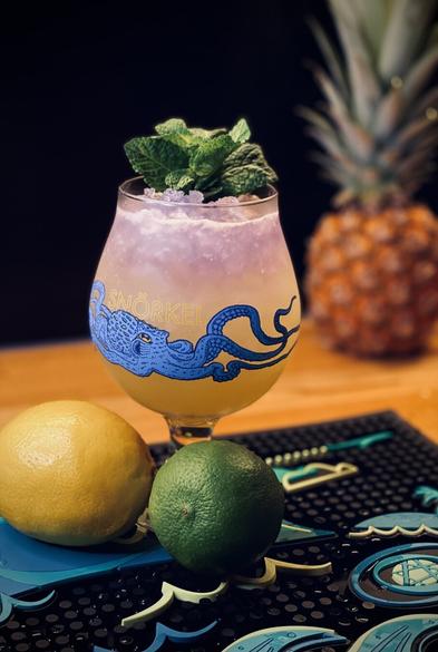 yellow cocktail with a purple float using Empress gin in a Jester King Snörkel glass garnished with a mint sprig sitting next to citrus fruit on a B-Rex bar mat