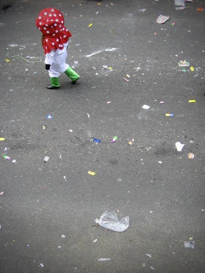 deutsch: das hochformatige bild zeigt von oben fotografiert den kleinen ausschnitt einer straße. zu sehen ist der nach einem karnevalsumzug mit bonbons und anderen sachen verschmutzte asphalt. in der linken oberen ecke des bildes ist ein verkleidetes kind zu sehen. es hat schwarze schuhe, eine grüne hose, ein weißes oberteil und eine rote kopfbedeckung mit weißen punkten. das kind bewegt sich nach links in richtung linke bildseite.

english: the portrait format picture shows a small section of a street photographed from above. you can see the asphalt soiled with sweets and other things after a carnival parade. in the top left corner of the picture you can see a child dressed up. it has black shoes, green trousers, a white top and a red headdress with white dots. the child is moving to the left towards the left side of the picture.