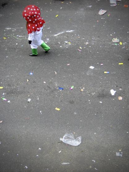deutsch: das hochformatige bild zeigt von oben fotografiert den kleinen ausschnitt einer straße. zu sehen ist der nach einem karnevalsumzug mit bonbons und anderen sachen verschmutzte asphalt. in der linken oberen ecke des bildes ist ein verkleidetes kind zu sehen. es hat schwarze schuhe, eine grüne hose, ein weißes oberteil und eine rote kopfbedeckung mit weißen punkten. das kind bewegt sich nach links in richtung linke bildseite.

english: the portrait format picture shows a small section of a street photographed from above. you can see the asphalt soiled with sweets and other things after a carnival parade. in the top left corner of the picture you can see a child dressed up. it has black shoes, green trousers, a white top and a red headdress with white dots. the child is moving to the left towards the left side of the picture.