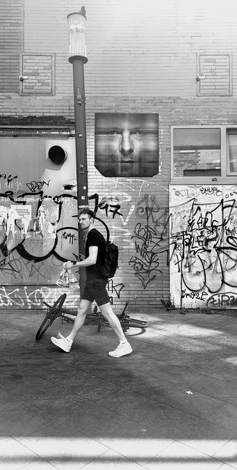 Black and white photo of a back walkway in Berlin, showing a young man in shorts looking left. A bicycle is lying on the floor. Above is an art image of David Bowie looking down. The walls are full of graffiti.the light is showing shadows in lines. The street lamp looks like it's leaning.