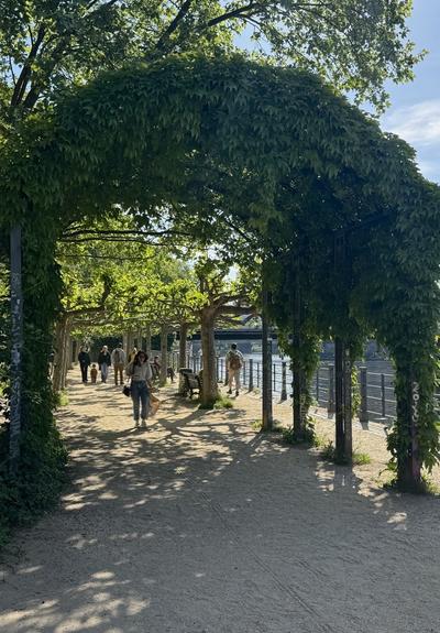 A shaded pathway with a leafy archway along a river, with people walking and sitting on benches.