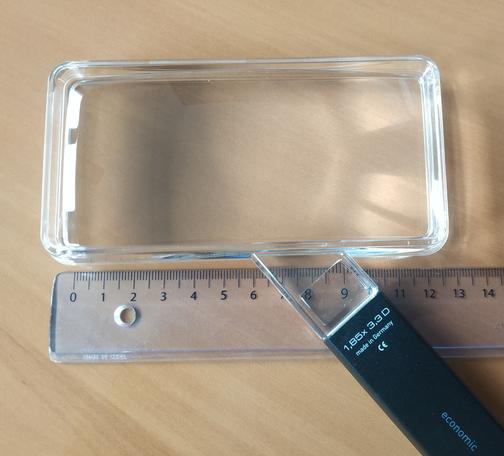Une loupe rectangulaire de 12 cm de longueur, posée sur une table. Une inscription sur le manche mentionne un grossissement de 1,85 et une vergence de 3,3 dioptries.