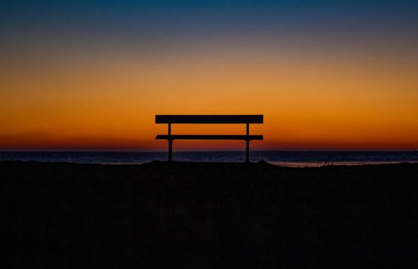 The silhouette of a park bench on a beach at sunset.